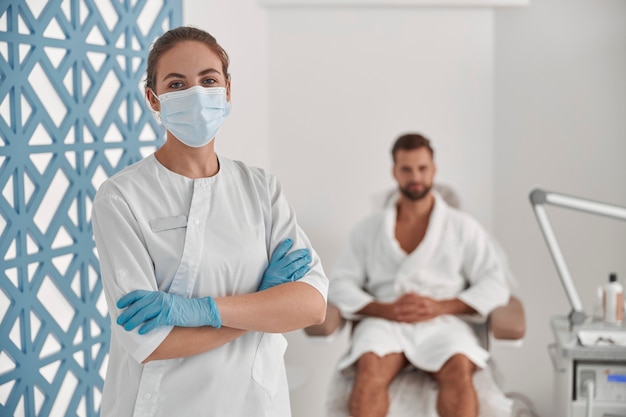 Esthéticienne en masque de protection et gants avec bras croisés près d'un patient homme assis dans un fauteuil