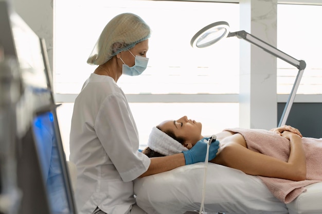 Esthéticienne en masque de protection et gants stériles à l'aide d'un stylo derma pendant la procédure de soin de la peau