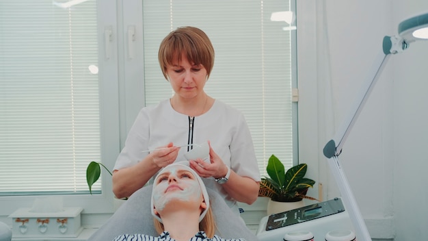 Photo esthéticienne mettant un masque crème sur le visage de la femme au salon de beauté des soins de la peau du visage