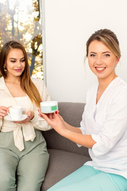 Esthéticienne offrant un produit pour la jeune femme tenant un pot en plastique blanc avec une crème assise sur le canapé