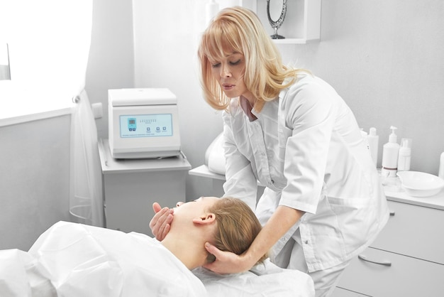 Une esthéticienne professionnelle donne à la jeune femme un massage manuel du visage et de la zone cervicale dans un salon de beauté blanc. Ã Â¡osmétologie, procédure de soins du visage