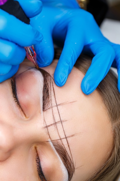 Esthéticienne tatouant les sourcils d'une femme à l'aide d'un équipement spécial pendant le maquillage permanent, gros plan.