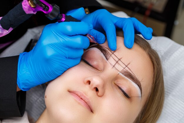 Esthéticienne tatouant les sourcils d'une femme à l'aide d'un équipement spécial pendant le maquillage permanent, gros plan.