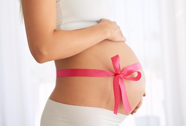 Estomac enceinte et femme avec un ruban rose dans la chambre à coucher pour l'annonce du sexe de la fille ou le cadeau de la fertilité Grossesse à la maison et future mère avec un nœud pour la douche de bébé santé maternelle ou ventre