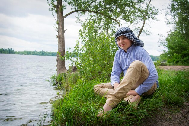 Photo en été, un beau garçon dans un foulard arabe est assis sur le rivage près de la rivière