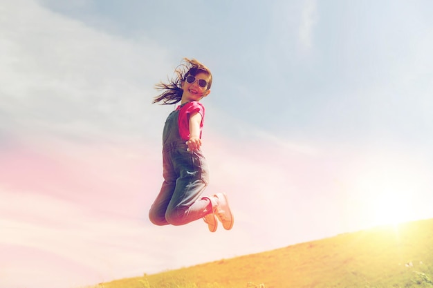 l'été, l'enfance, les loisirs et les gens concept - petite fille heureuse sautant haut sur le champ vert et le ciel bleu à l'extérieur