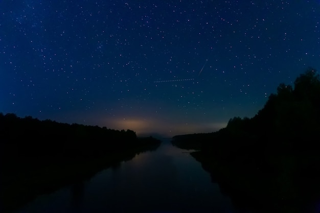 Les étoiles décolorées dans le ciel du petit matin se reflètent dans une rivière calme. La trace d'un avion en vol croise la trace d'un astéroïde en chute libre dans le ciel nocturne.