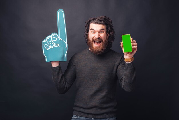 Étonné crier jeune homme barbu avec un gant de ventilateur montrant un écran vert sur le téléphone