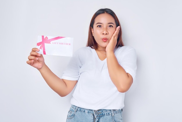 Étonné jeune femme blonde fille asiatique portant un t-shirt blanc décontracté tenant un bon de coupon de certificat-cadeau touchant la joue avec la main isolé sur fond blanc concept de mode de vie des gens