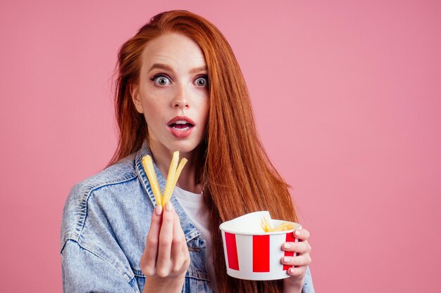 Photo Étonné et surpris jolie femme gingembre aux cheveux longs rouges foxy tenant un seau de boîte en carton d'emballage avec des frites en studio rose.concept de régime.