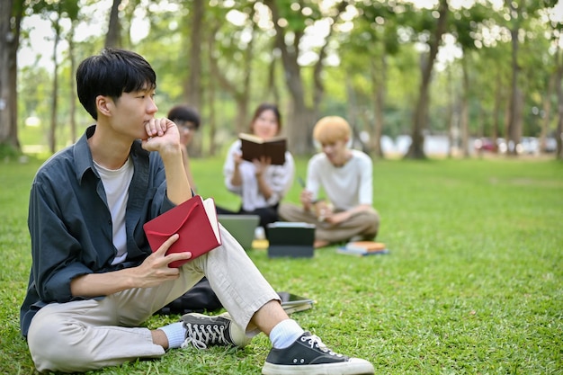 Un étudiant asiatique réfléchi est assis sur l'herbe dans le parc du campus avec son livre