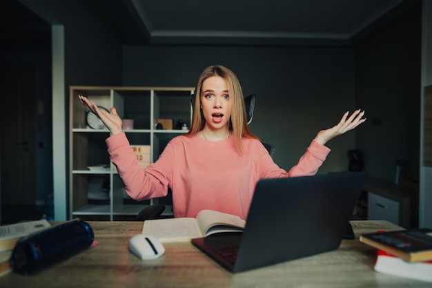 Un étudiant confus dans un pull rose est assis à la maison sur un ordinateur portable tout en faisant des devoirs universitaires