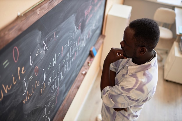 Étudiant ou enseignant afro-américain pensif debout devant un tableau noir avec des formules et des équations écrites en classe