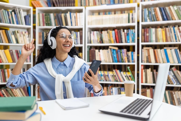 Photo un étudiant joyeux écoute de la musique sur des écouteurs à l'aide d'un smartphone dans une bibliothèque entourée de