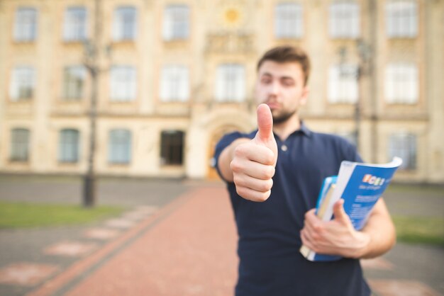 Un étudiant avec des livres à la main se tient sur le campus de l'université et montre un coup de pouce.