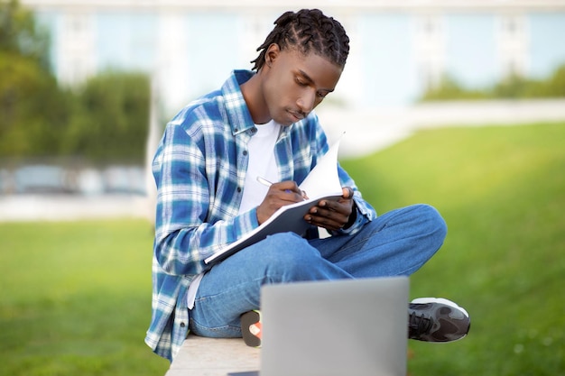 Un étudiant noir étudie à l'extérieur, écrit et navigue sur son ordinateur portable.