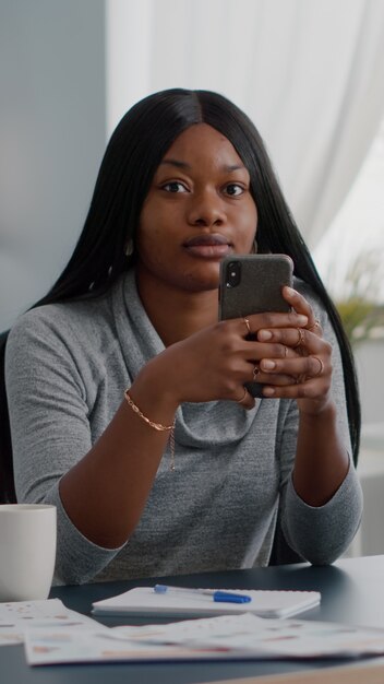 Étudiant Noir Tenant Un Téléphone Dans Les Mains Discutant Avec Des Personnes Parcourant Des Informations De Communication Assises Au Bureau Dans Le Salon. Adolescent Regardant Sur Les Médias Sociaux Partageant Des Conseils De Style De Vie Avec Des Amis Du Groupe