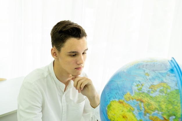 Un étudiant réfléchi dans une chemise blanche est assis à une table devant un globe Le concept de voyage leçon de géographie