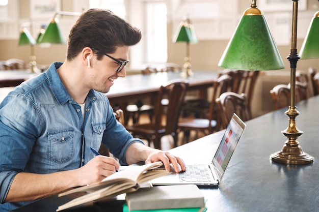 Étudiant De Sexe Masculin Souriant Qui étudie à La Bibliothèque, à L'aide D'un Ordinateur Portable