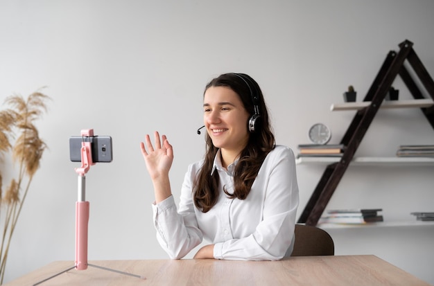 Un étudiant souriant avec un casque sans fil suit une leçon en ligne depuis chez lui via un smartphone debout sur un trépied