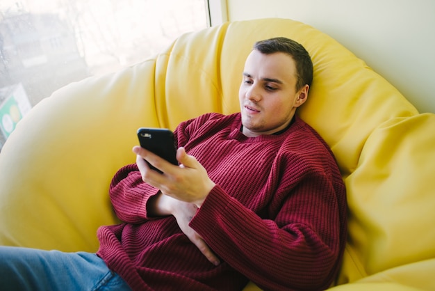 Un étudiant souriant se repose pendant une pause dans un sac de fauteuil jaune et utilise un smartphone. Loisirs passifs.