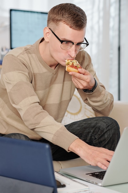 Étudiant à L'université Dans Des Verres Mangeant Une Tranche De Pizza En Travaillant Sur Ordinateur Portable à La Maison