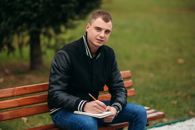 Un étudiant en veste noire est assis dans un parc sur un banc et écrit ses pensées dans un cahier