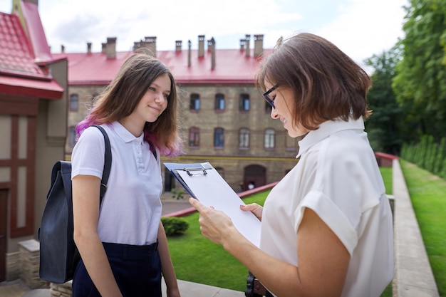 Une étudiante adolescente parle avec un enseignant, une femme avec un presse-papiers lisant et écrivant dans un document papier de l'extérieur de l'école. Retour à l'école, retour au collège, concept d'éducation