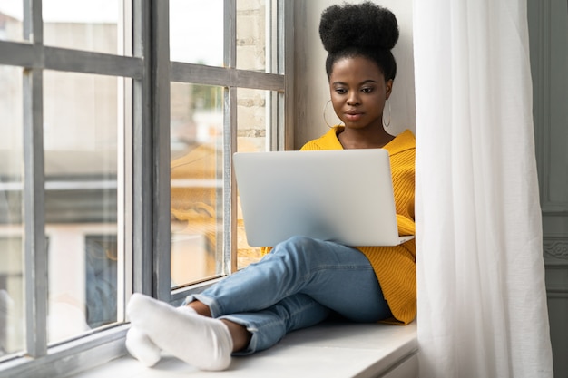 Une étudiante afro-américaine avec une coiffure afro porte un cardigan jaune, assise sur le rebord de la fenêtre, travaillant à distance sur un ordinateur portable, apprenant à l'aide d'un cours en ligne. Auto-éducation, préparation à un examen.