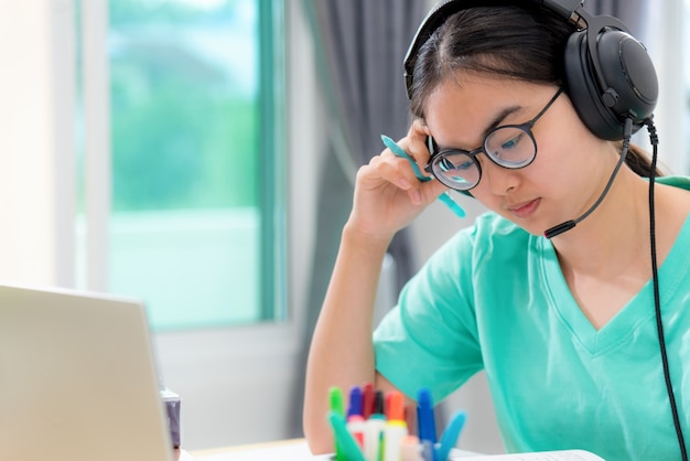 Photo Étudiante asiatique adolescente avec des lunettes casque assis à la lecture sérieuse d'un livre s'inquiéter à l'aide d'un ordinateur portable sur la table apprenant l'étude en ligne. éducation d'une classe d'université à la maison