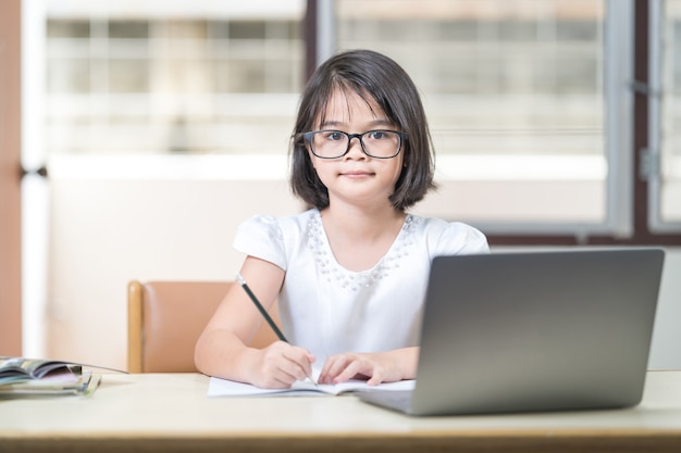 Une étudiante asiatique avec des lunettes étudie en ligne, fait ses devoirs sur un ordinateur portable à la maison. Éducation, concept Banque de Photo