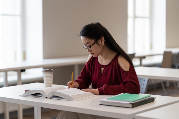 Une étudiante asiatique prend des notes à partir d'un manuel faisant ses devoirs à la bibliothèque s'asseoir au bureau de l'école