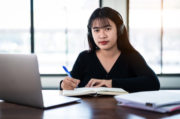 Une étudiante asiatique souriante porte des écouteurs sans fil écrivez sur le cahier pour étudier la langue en ligne, regardez et écoutez le conférencier, webinaire par appel vidéo e-learning à la maison, enseignement à distance