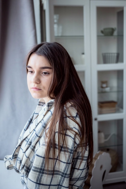 Étudiante aux cheveux bruns réfléchie en tenue décontractée regardant loin tout en se tenant près de la fenêtre à la maison