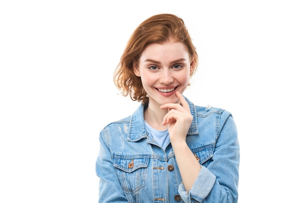 Une étudiante aux cheveux roux pense joyeusement et regarde la caméra tout en tenant son doigt près de son menton. Femme réfléchie pensive dans une veste en jean bleu sur fond blanc. Espace de copie