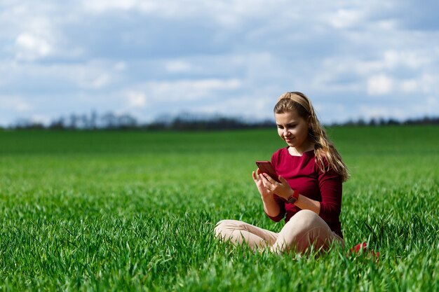 Étudiante De La Belle Jeune Femme Avec Un Téléphone Dans Les Mains Assis Sur L'herbe. La Fille Prend Des Selfies Et Prend Des Photos De Selfie. Elle Sourit Et Profite D'une Journée Chaude. Photo Conceptuelle Sur Smartphone