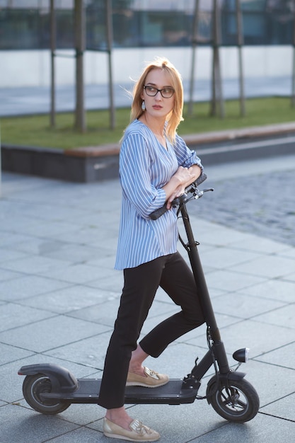 Une étudiante blonde et pensive à lunettes conduit son nouveau scooter électro noir solide à l'université.