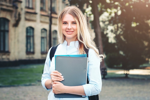 Une étudiante blonde sourit et tient un dossier et un cahier à la main sur un fond universitaire