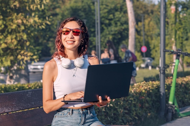 Une étudiante avec un casque travaille et étudie sur un ordinateur portable assis sur le banc dans le parc
