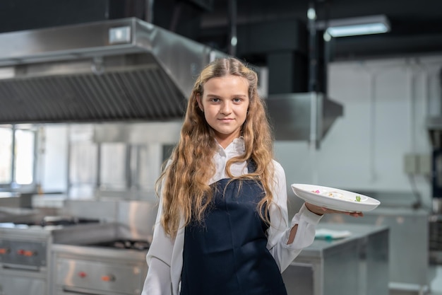 Une étudiante chef junior portant un tablier tenant son apéritif dans un plat tout en étudiant la cuisine