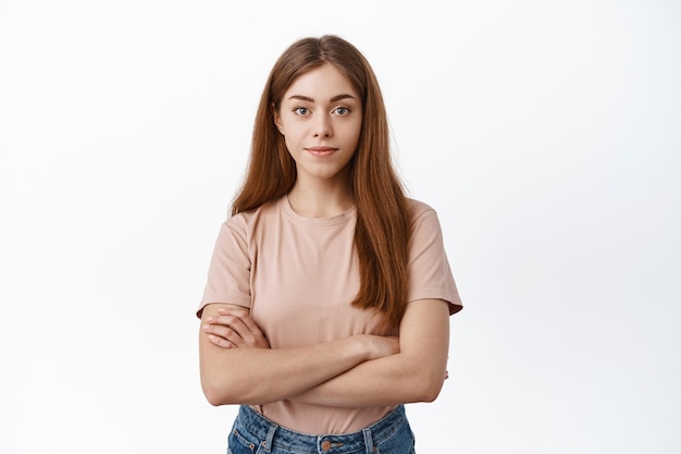 Une étudiante croise les bras sur la poitrine et sourit à l'avant, a l'air déterminée et sûre d'elle, se tient contre un mur blanc