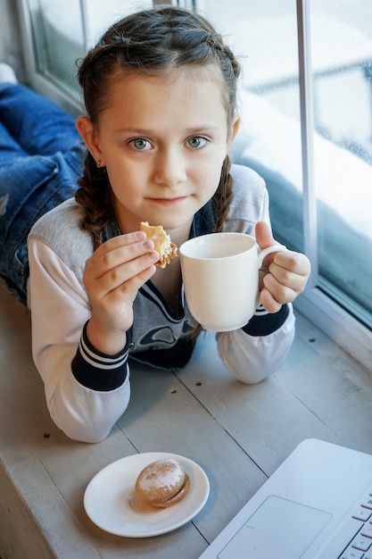 Une étudiante étudie en ligne avec un professeur d'appel vidéo, une jeune fille heureuse écoute une conférence sur un ordinateur portable à la maison et boit du thé ? manger des biscuits. Coronavirus covid-19.