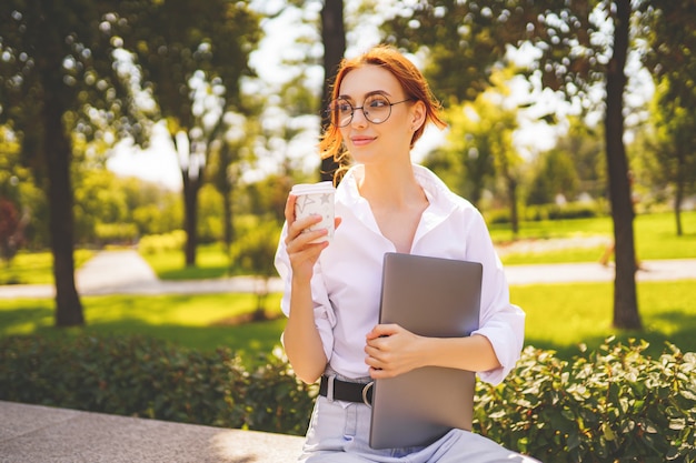 Une étudiante heureuse dans des lunettes et une chemise blanche tient un ordinateur portable et un café assis dans le parc de la ville