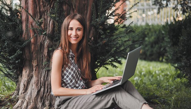 Une étudiante joyeuse est assise sur l'herbe près de l'arbre avec un ordinateur portable sur ses genoux et compose le clavier