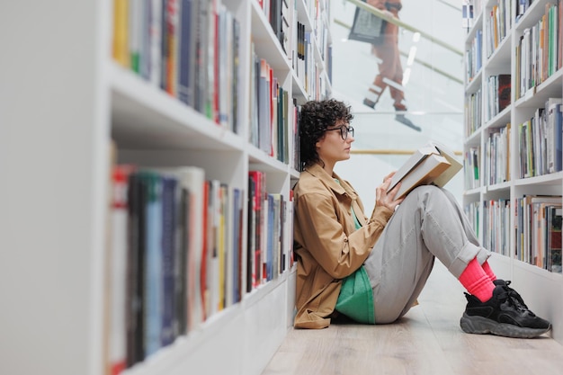 Une étudiante lit un livre assis parmi les étagères de la bibliothèque ou du coworking