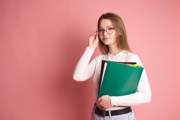 Une étudiante mignonne avec des lunettes tient des dossiers colorés sur fond rose