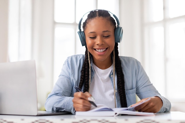 Une étudiante noire heureuse qui étudie avec des écouteurs et un ordinateur portable.