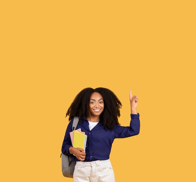 Une étudiante noire heureuse avec un sac à dos et des cahiers pointant vers le haut dans l'espace libre sur le jaune