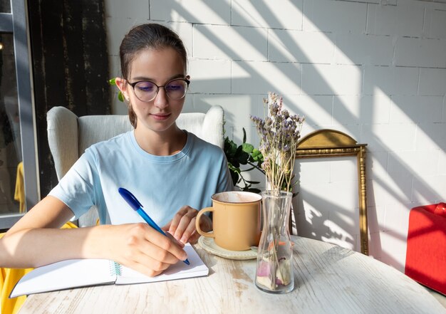 Une étudiante pensive écrit avec un stylo un devoir dans un cahier. Portrait d'une jeune fille brune caucasienne à lunettes et un chemisier bleu dans un café avec des ombres diagonales. Concept de lecture de livres papier