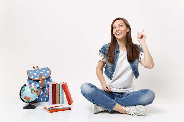 Étudiante Pensive Souriante En Vêtements En Denim Rêvant, Pointant L'index Vers Le Haut, Assise Près Du Globe, Sac à Dos, Livres Scolaires Isolés